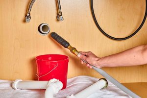 Foreground Tools: Various plumbing tools are scattered on the floor. These include wrenches, pipes, and connectors. A white U-shaped pipe is prominently visible on the left side of the image. Metallic pipes and flexible hoses are among the tools. Person at Work: A person wearing blue gloves is actively working near a white toilet. The toilet is partially visible on the right side of the image. It seems like the person is either fixing an existing plumbing issue or installing new plumbing components. Background: The floor features square tiles, and there’s a tiled wall in the background with a simple pattern. Overall, this image captures an ongoing plumbing maintenance and Gas Line Installation Denton TX